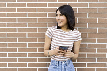 Smiling young woman holding mobile phone while leaning on brick wall - GIOF13040