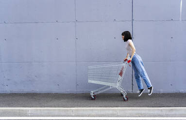 Woman with short hair playing with shopping cart on footpath - GIOF13058