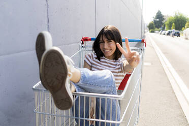 Smiling woman gesturing while sitting in shopping cart on footpath - GIOF13063