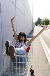 Carefree woman with arms raised sitting in shopping cart - GIOF13064