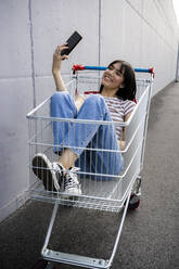 Smiling young woman taking selfie through smart phone while sitting in shopping cart - GIOF13067