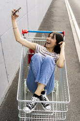 Cheerful woman taking selfie through smart phone in shopping cart - GIOF13068