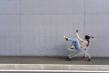 Junge Frau macht Selfie mit Smartphone im Einkaufswagen auf dem Gehweg - GIOF13069