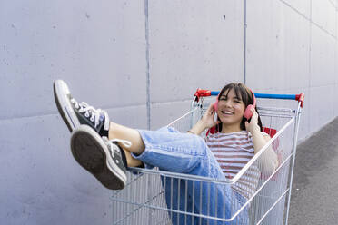 Lächelnde Frau hört Musik über Kopfhörer, während sie mit hochgelegten Füßen im Einkaufswagen sitzt - GIOF13071