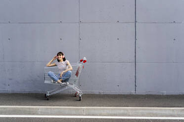 Smiling woman listening music through headphones while sitting cross-legged in shopping cart - GIOF13075