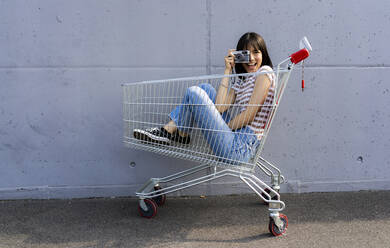 Cheerful woman photographing through camera in shopping cart - GIOF13078