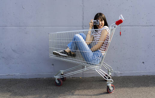 Fröhliche Frau, die durch eine Kamera im Einkaufswagen fotografiert - GIOF13078