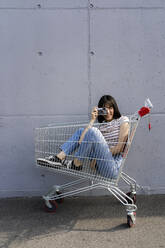 Woman photographing through camera while sitting in shopping cart - GIOF13079