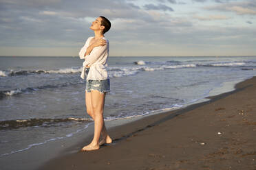 Young woman hugging herself while standing at beach - VEGF04746