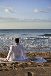 Frau schaut auf das Meer, während sie auf einer Decke sitzt - VEGF04754