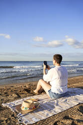 Frau fotografiert am Strand sitzend das Meer mit dem Handy - VEGF04756