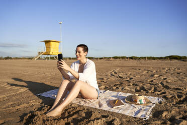 Lächelnde Frau, die ein Mobiltelefon benutzt, während sie am Strand sitzt - VEGF04757