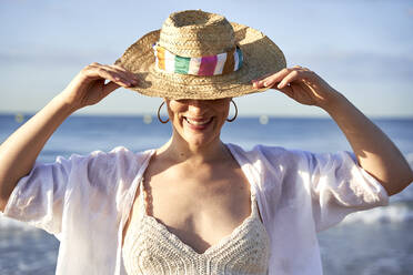 Frau lächelt und hält Hut am Strand - VEGF04760
