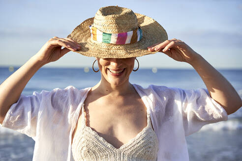 Woman smiling while holding hat at beach - VEGF04760