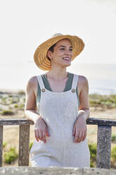 Smiling young woman wearing hat leaning on railing - VEGF04764