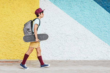 Boy with backpack and skateboard walking by multi colored wall - JCMF02136