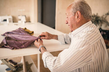Senior male tailor examining fabric working while sitting at workbench - GRCF00768