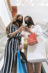 Freundinnen mit Gesichtsschutz tragen Einkaufstaschen, während sie in einem Einkaufszentrum ihr Smartphone benutzen - JRVF01218