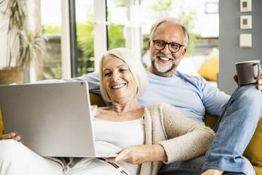 Frau lächelt bei Videoanruf über Laptop, während sie mit einem Mann auf dem Sofa sitzt - UUF24074