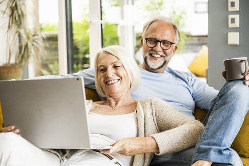 Woman smiling at video call through laptop while sitting with man on sofa - UUF24074