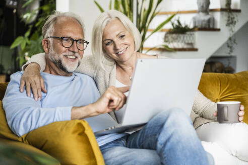 Lächelnder reifer Mann, der einen Laptop benutzt, während er mit einer Frau auf dem Sofa zu Hause sitzt - UUF24078