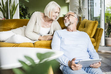 Mature man with digital tablet looking at woman in living room at home - UUF24087