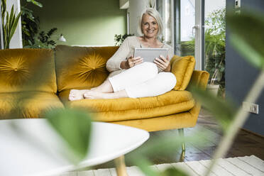 Smiling woman holding digital tablet while sitting on sofa - UUF24090