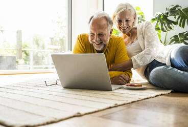 Smiling couple using laptop while relaxing on carpet at home - UUF24104