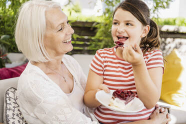 Enkelin isst Kuchen, während sie mit ihrer Großmutter auf dem Balkon sitzt - UUF24116