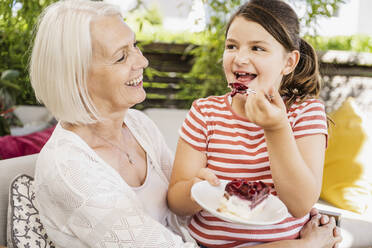 Granddaughter eating cake while sitting with grandmother at balcony - UUF24116