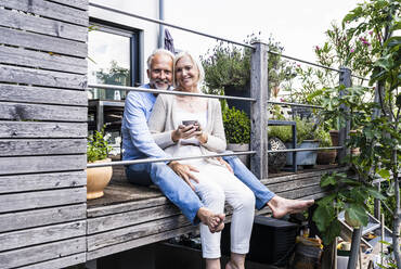 Smiling man sitting with woman holding mobile phone at balcony - UUF24139