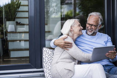 Frau umarmt Mann mit digitalem Tablet, während sie auf dem Balkon sitzt - UUF24143