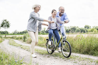 Gut gelaunte Großeltern helfen ihrer Enkelin beim Fahrradfahren auf einem unbefestigten Weg - UUF24156