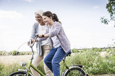 Grandmother helping granddaughter while riding bicycle at meadow - UUF24162