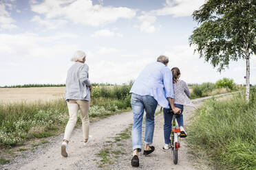 Enkelin fährt Fahrrad, während die Großeltern auf einem unbefestigten Weg laufen - UUF24166