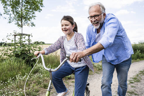 Fröhlicher Großvater spielt mit seiner Enkelin, die auf einem unbefestigten Weg Rad fährt - UUF24167