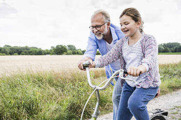 Smiling mature man assisting girl riding bicycle at meadow - UUF24168