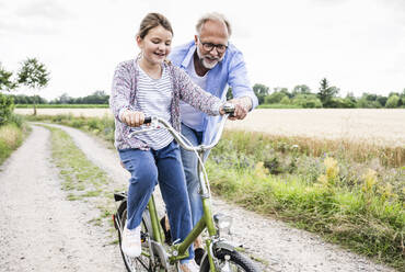 Grandfather teaching cycling to granddaughter on dirt road - UUF24169