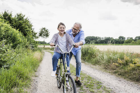 Großvater hilft seiner Enkelin beim Fahrradfahren auf einem unbefestigten Weg - UUF24170