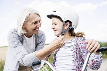 Grandmother putting cycling helmet to girl standing with bicycle - UUF24171