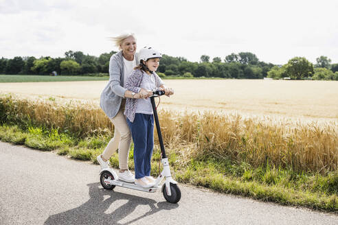 Happy granddaughter and grandmother playing while riding push scooter during sunny day - UUF24182