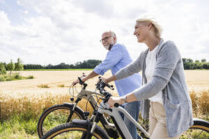 Smiling couple with bicycles walking by field during sunny day - UUF24191