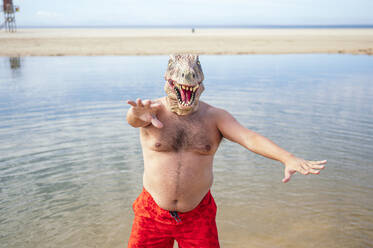 Shirtless man wearing lizard mask gesturing at beach - JCMF02173