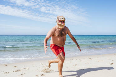 Mann mit Eidechsenmaske läuft an einem sonnigen Tag am Strand entlang - JCMF02177