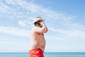 Älterer übergewichtiger Mann, der im Urlaub am Strand mit seinem Smartphone telefoniert - JCMF02179