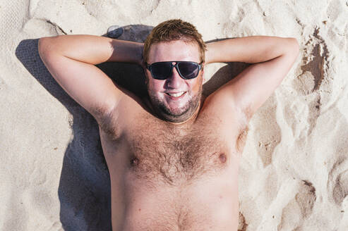Lächelnder schwuler Mann, der sich an einem sonnigen Tag im Sand entspannt - JCMF02184