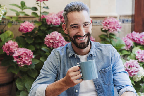 Bärtiger Geschäftsmann mit Kaffee vor blühenden Pflanzen im Hinterhof - EBBF04356