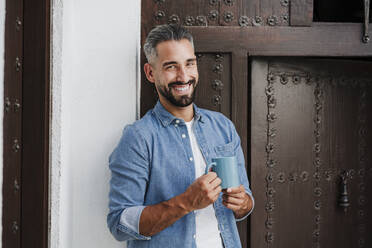 Gut aussehender Geschäftsmann, der eine Kaffeetasse hält, während er vor einer Holztür steht - EBBF04358