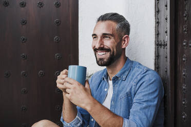 Smiling male freelancer looking away while holding coffee mug by door - EBBF04362