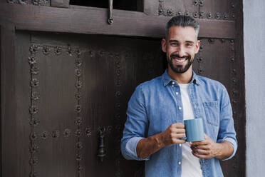Geschäftsmann lächelt, während er einen Kaffeebecher vor einer Tür hält - EBBF04363